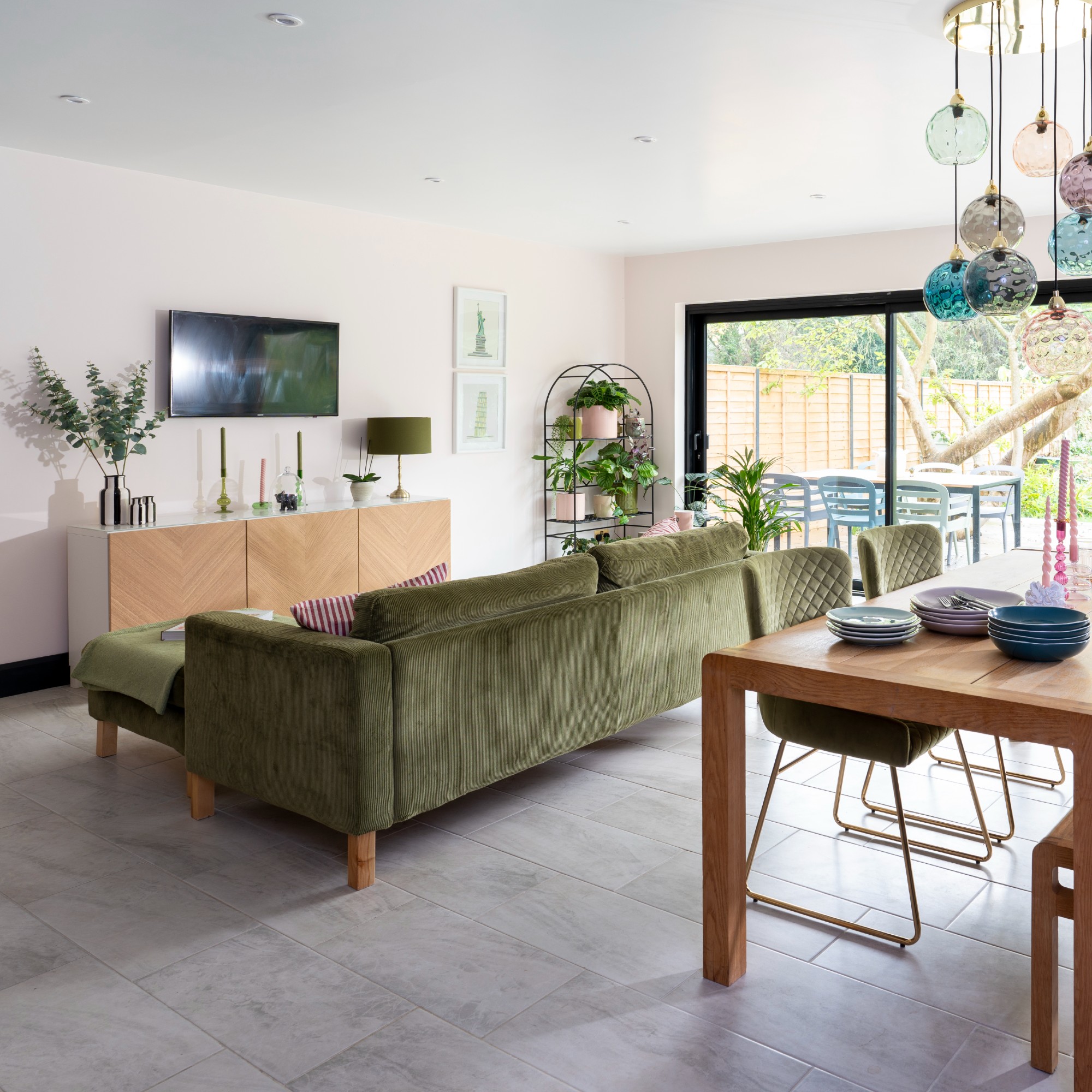 An open-plan living room with an L-shaped green velvet sofa and a sideboard displaying decorative items and a TV hung above it