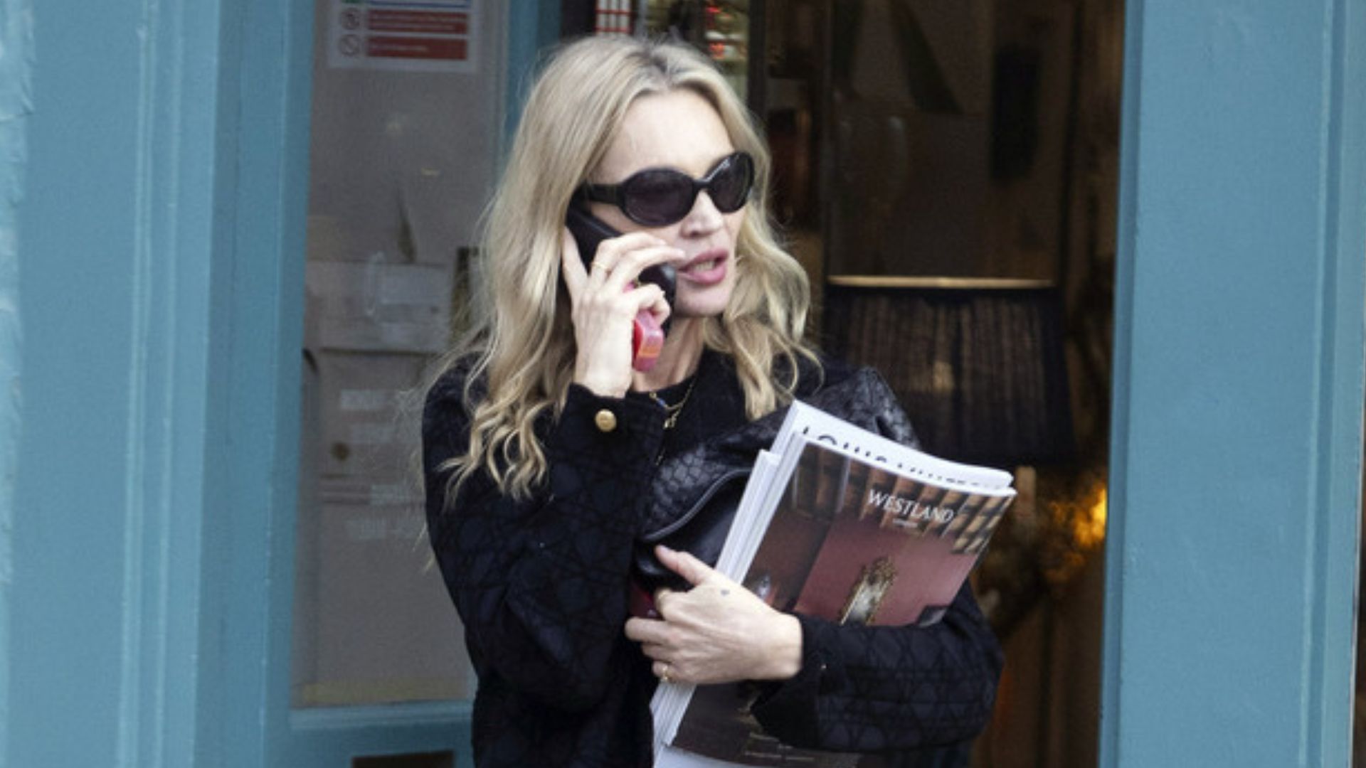Kate Moss steps out, taking a phone call. She wears blue bootcut jeans with a black jumper, black loafers and a black belt. She carries a stack of magazines in the crook of her arm.