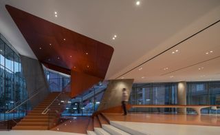 Interior view of the new Medical Center for New York’s Columbia University. Spacious atrium in white muted orange and reddish brown colors. Glass windows surround the area, and there is a staircase to the left that leads to the upper floors.