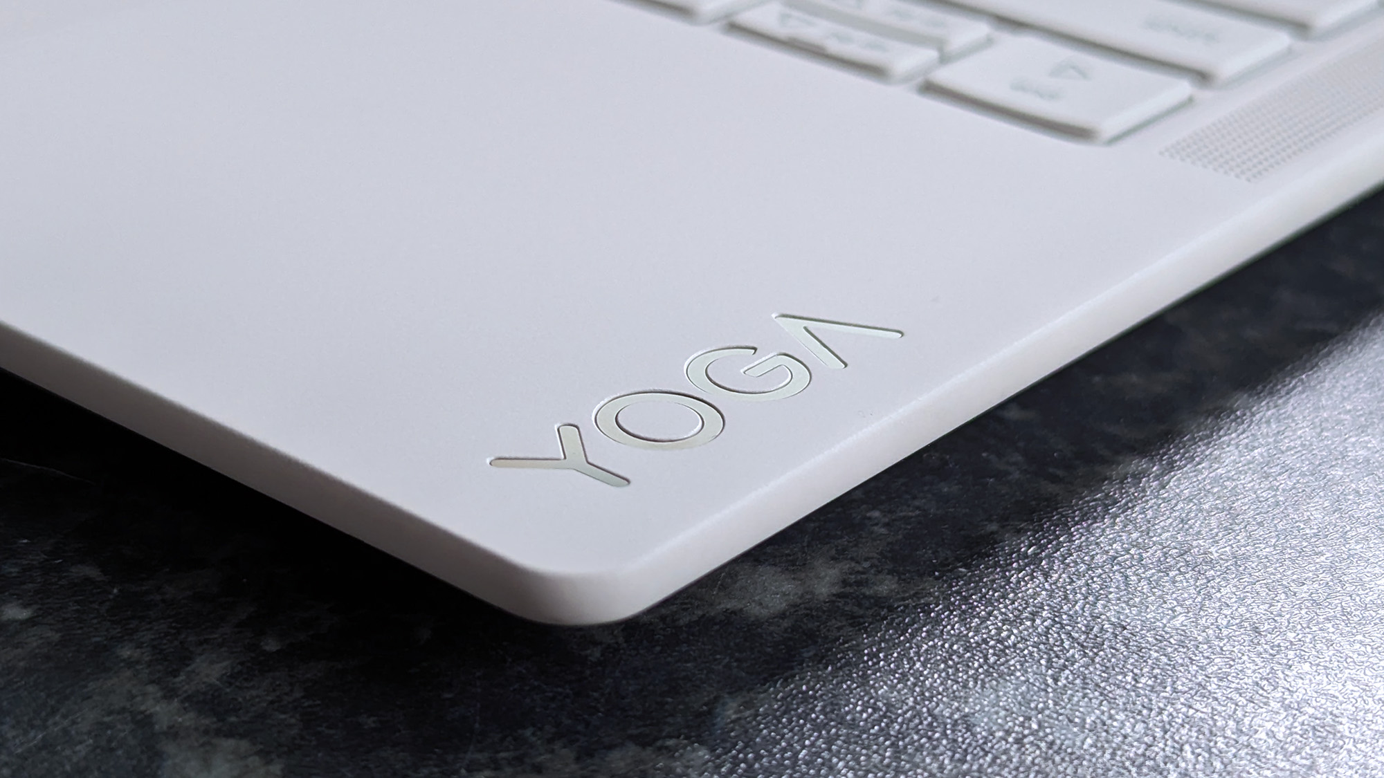 Close-up of a Lenovo Yoga Slim 7i Ultra (Gen 11) Aura Edition laptop corner with the word "YOGA" engraved in sleek letters. The laptop rests on a speckled dark surface.