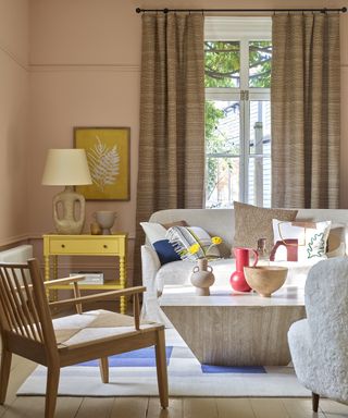 A pale pink living room with neutral wooden furniture and pops of primary colors.