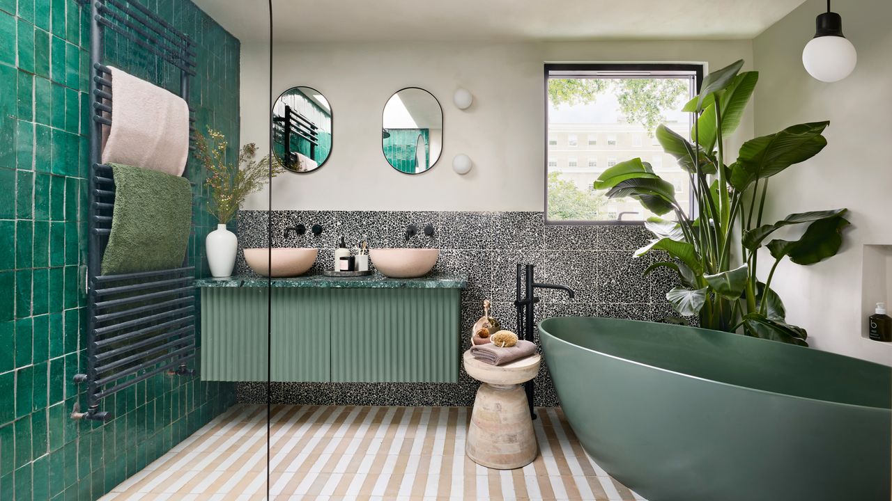 bathroom with beige and white striped floor tiles, a large green bathtub, an oversized banana plant and a green fluted bathroom console unit with twin pink/beige basins and black hardware beside a shower area with emerald green tiles a clear glass shower panel and black hardware