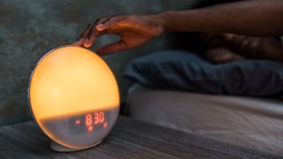 A hand turning off a sunrise alarm clock on a bedside table in a darkened room