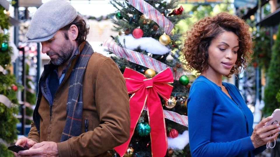 A woman and a man stand in front of many christmas trees looking at their phones and not realizing they&#039;re texting each other!