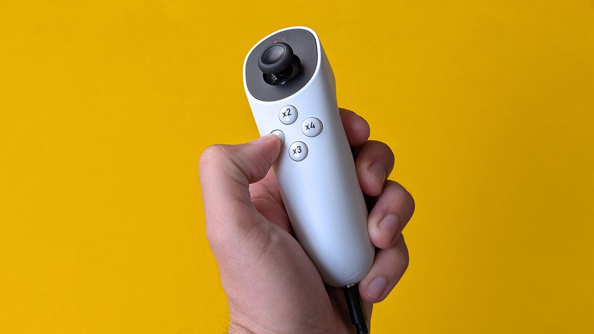 A hand holds a white XBOX Adaptive Joystick featuring a black thumbstick and four round buttons. The device is connected by a cable at the bottom and set against a bright yellow background.
