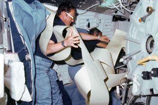 Two astronauts wrangle a spool of computer printouts inside the space shuttle.