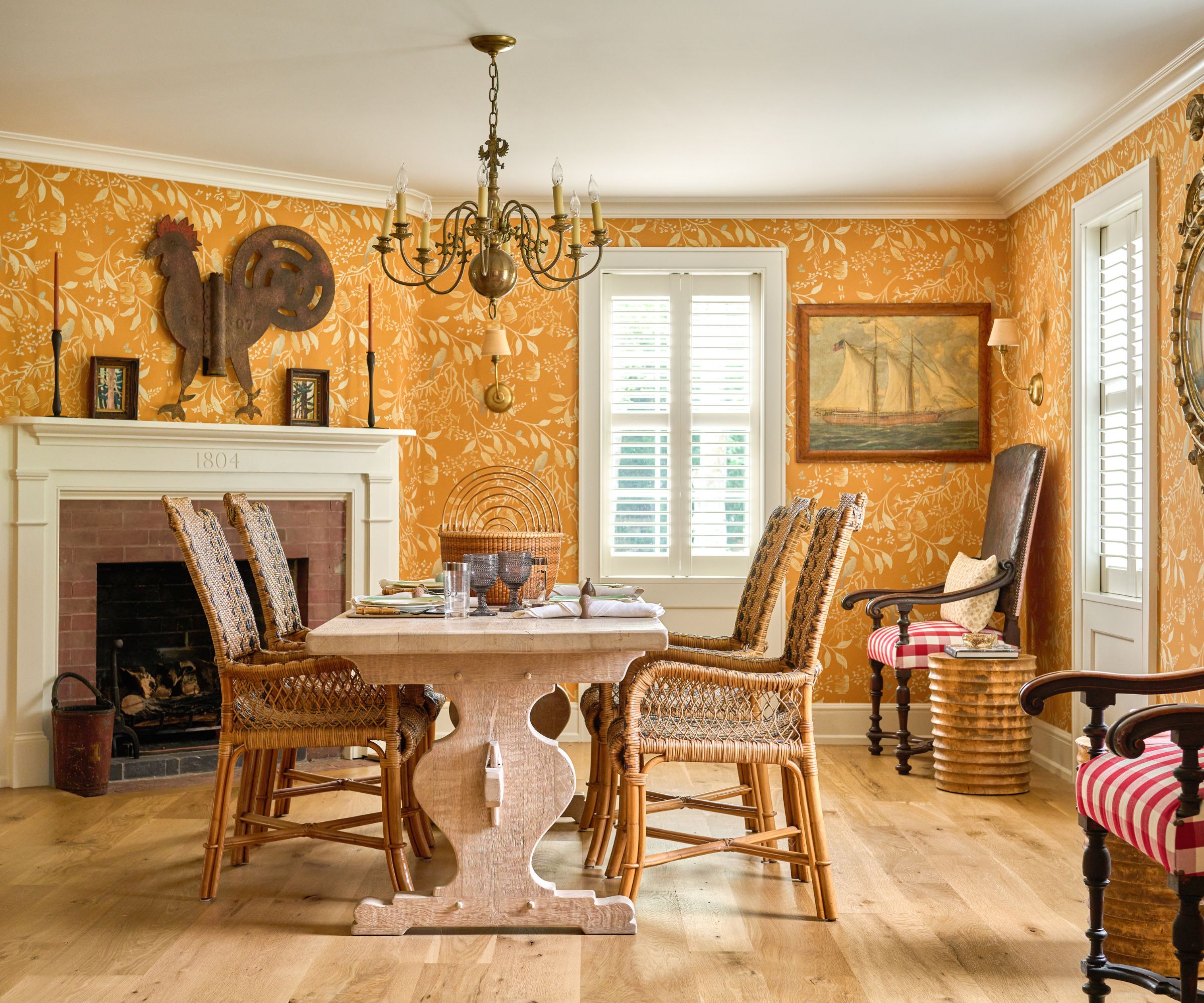 Dining room with yellow patterned wallpaper wooden table, rattan chairs and a white fireplace
