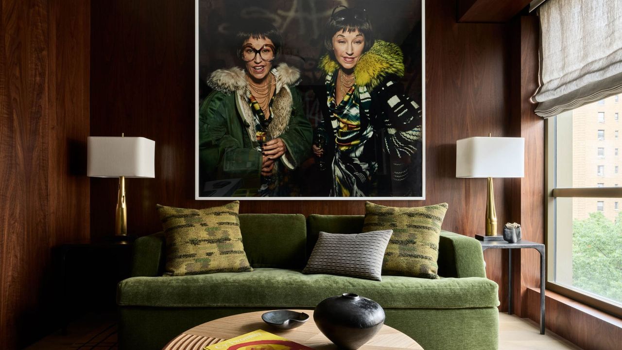 A wood living room with a large framed picture a pair of white lamps with gold stands, an olive green sofa, textured pillows, a round wood coffee table, a black vase, and a coffee table book