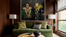 A wood living room with a large framed picture a pair of white lamps with gold stands, an olive green sofa, textured pillows, a round wood coffee table, a black vase, and a coffee table book