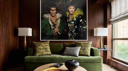 A wood living room with a large framed picture a pair of white lamps with gold stands, an olive green sofa, textured pillows, a round wood coffee table, a black vase, and a coffee table book