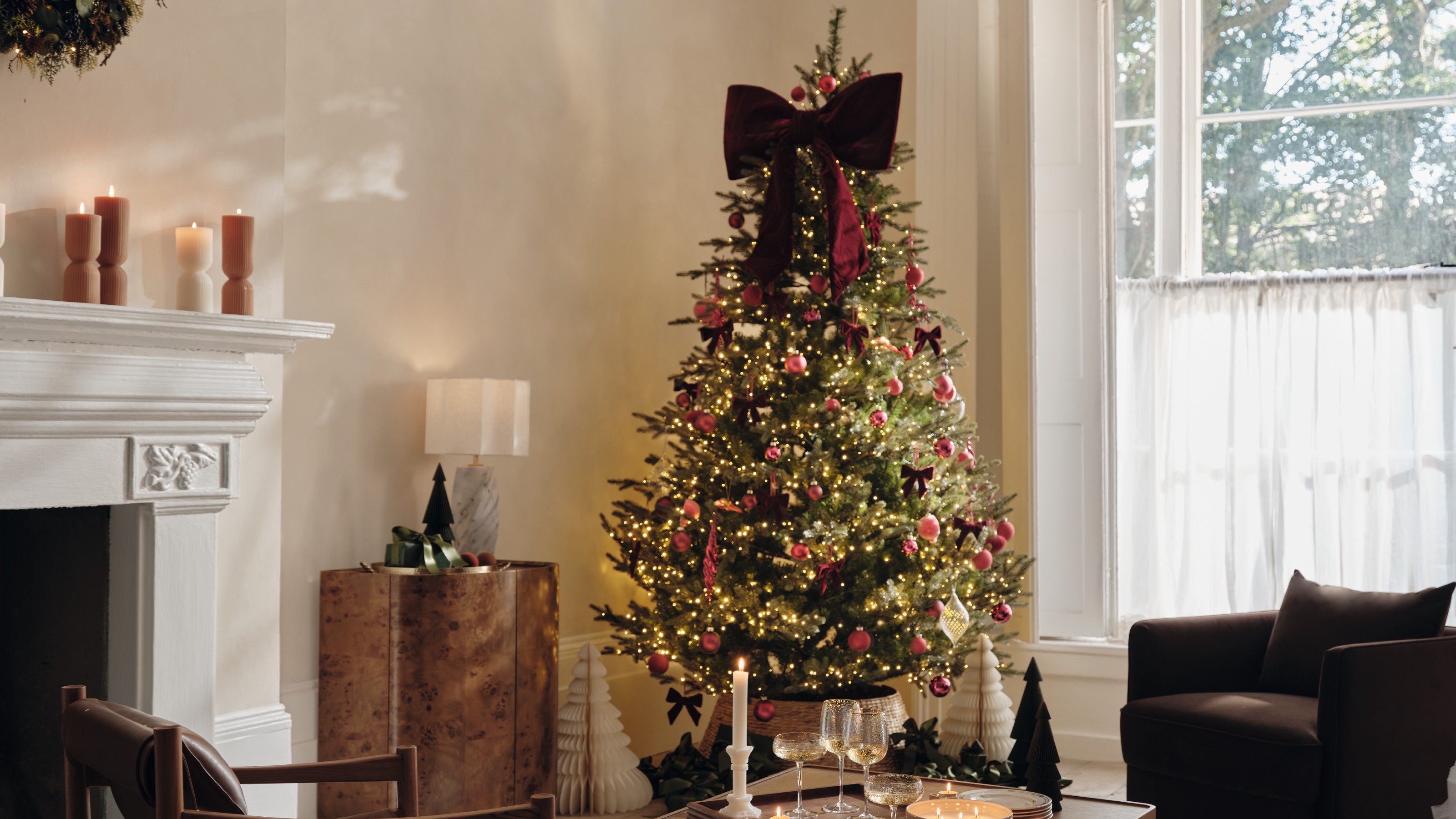 A modern living room decorated for Christmas with a lit-up Christmas tree in the corner decorated with a large red velvet bow