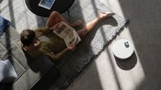 Top-down photograph of person holding magazine sat on gray rug in gray, neutral living room, pictured next to a white Dreame robot vacuum cleaner. Sunbeams are shining light across the floor.