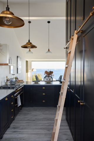 navy kitchen with cabinets up to ceiling with ladder, view to sea, copper pendant lights, galley style, white countertops, artwork, gray floor