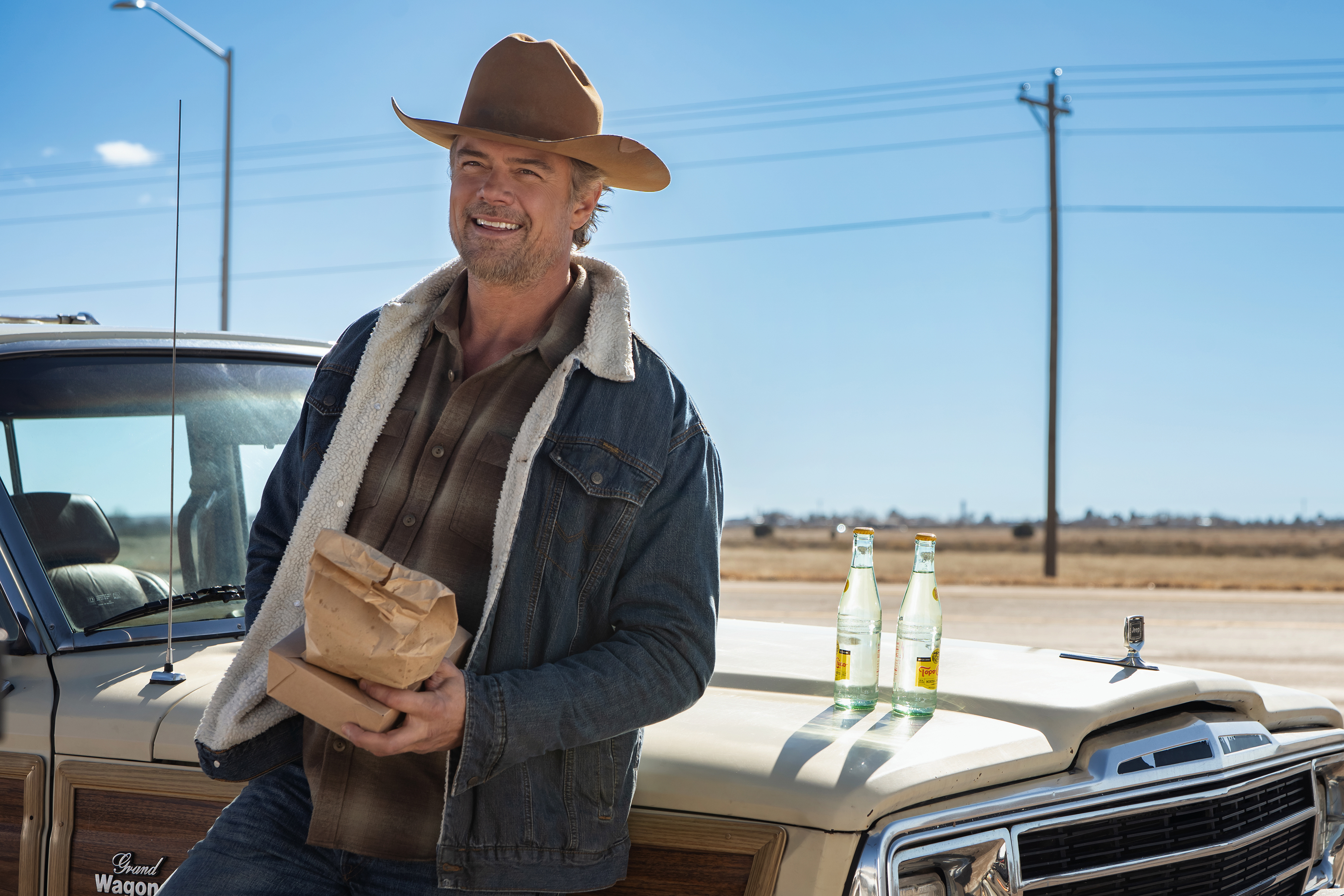 Josh Duhamel as Staten smiling and drinking beers while leaning against his car on a highway in a still from ransom canyon season 2