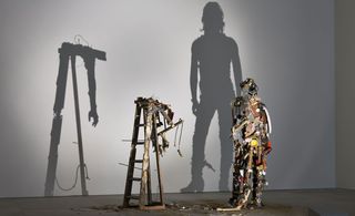 A closer view of one shadow sculpture made out of junk, metal, and wood. The light shines on the sculpture and casts a shadow on the wall. The shadow portrays a woman to the right, and the arm of a man turned towards the floor.