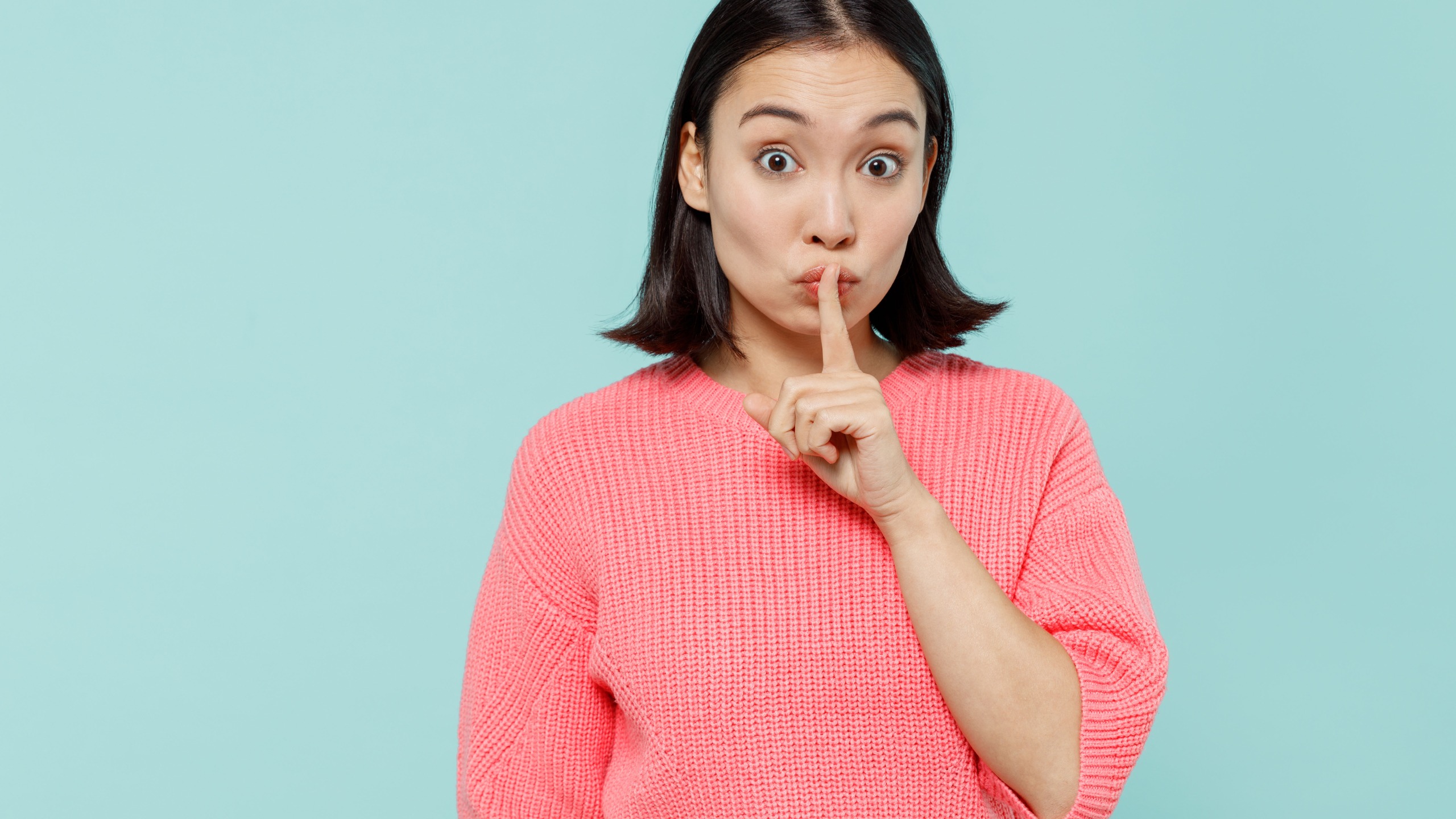 Young secret woman of Asian ethnicity 20s in pink sweater say hush be quiet with finger on lips shhh gesture.