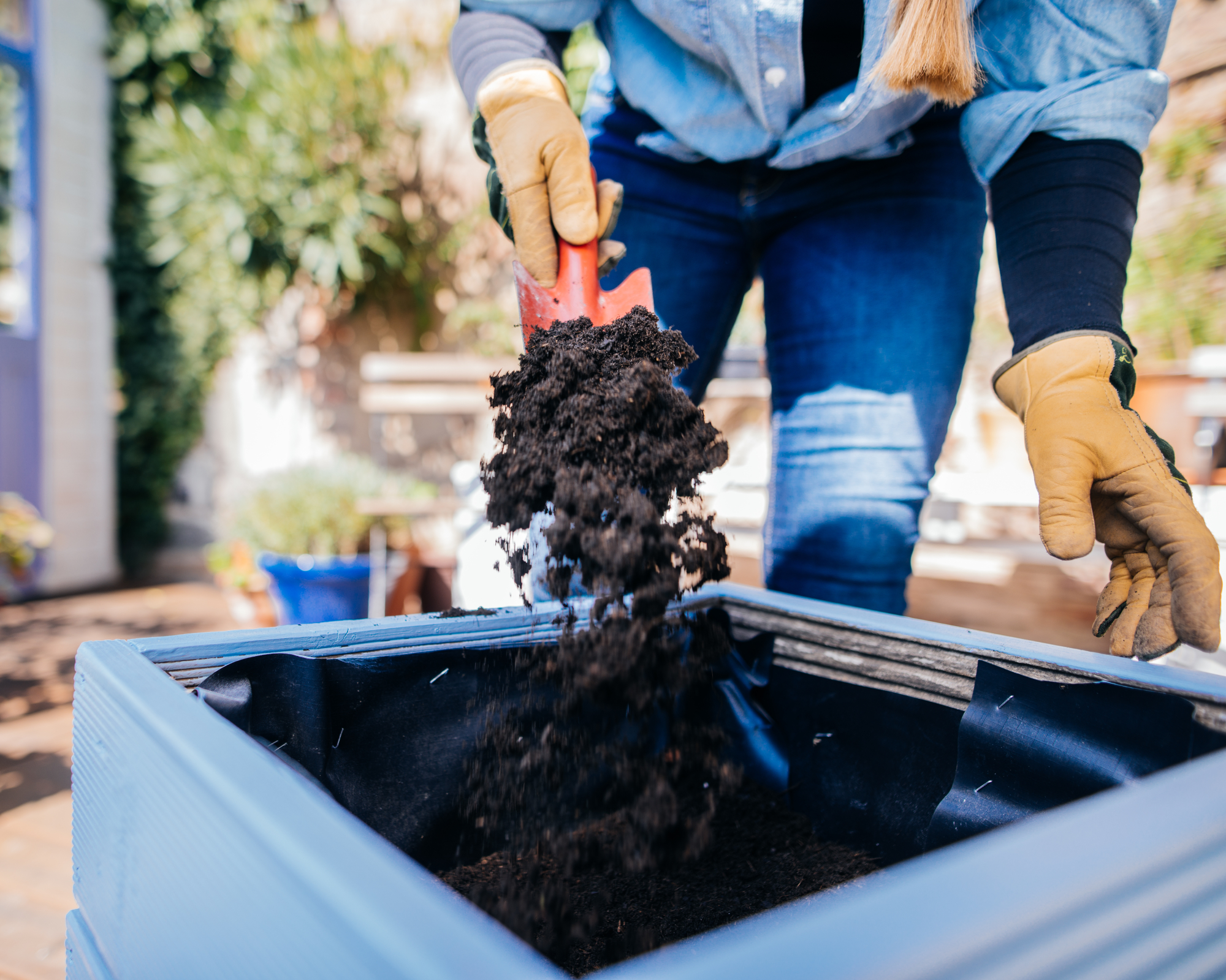 woman using a red trowel to fill a large container with potting soil on a patio