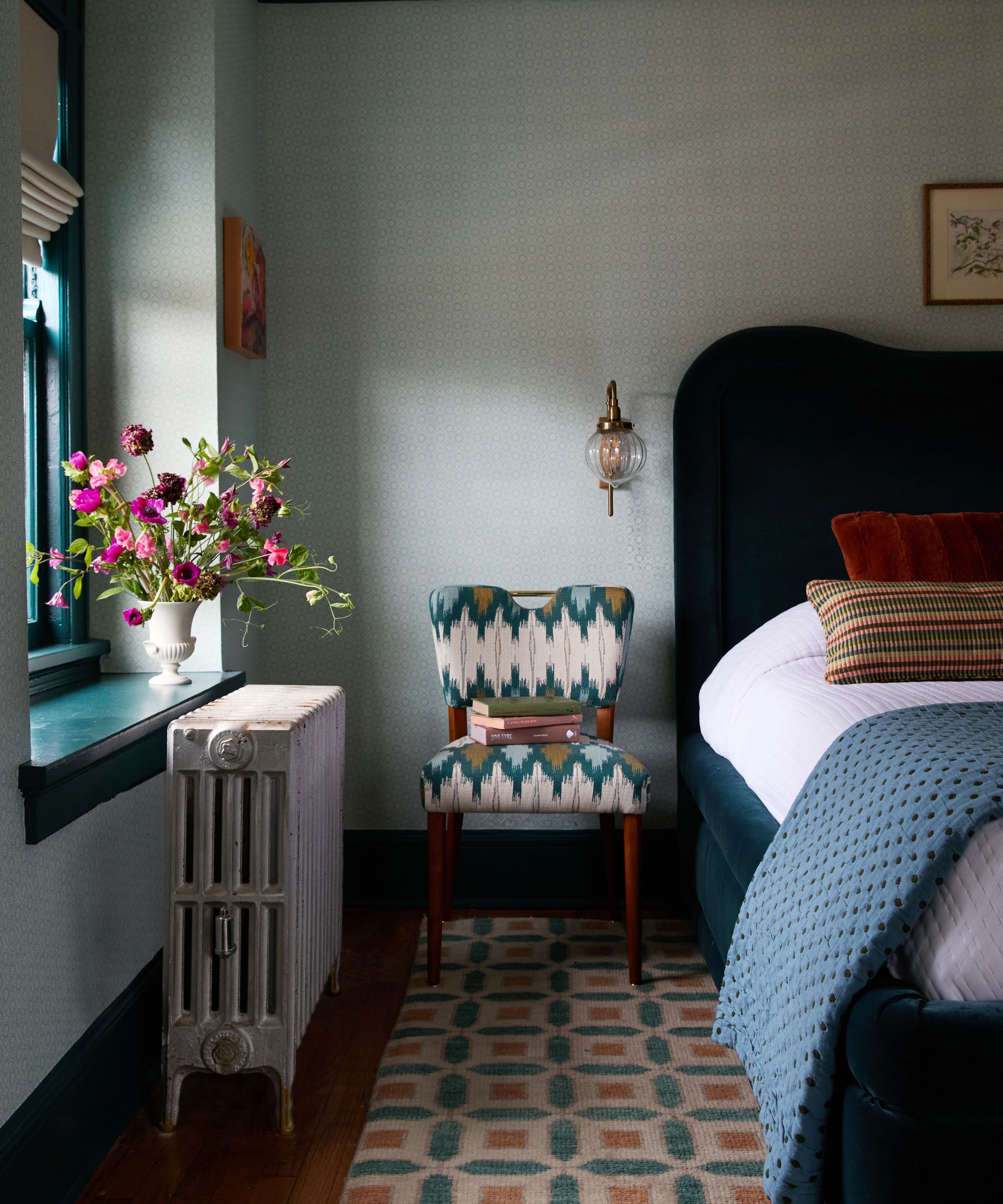 A bedroom with neutral walls, a bed with a teal velvet headboard and a geometric print rug, and a patterned teal accent chair.