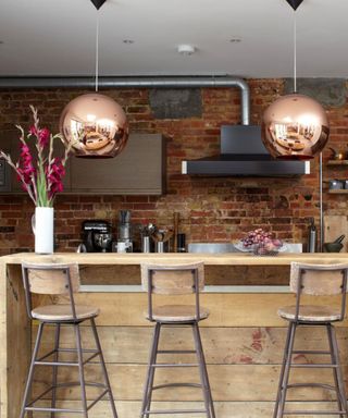 An industrial kitchen with a light wooden kitchen island with three wooden chairs in front of it, two copper pendant lights above it, and a brick wall behind it