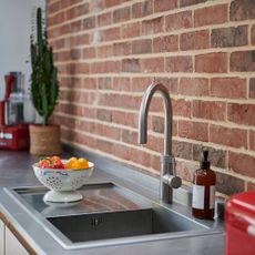 a kitchen with a brick wall behind a silver kitchen sink with a white colander containing sweet peppers resting on the side, and a catci in a pot at the far end of the bench next to a red food processor