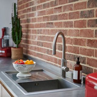 a kitchen with a brick wall behind a silver kitchen sink with a white colander containing sweet peppers resting on the side, and a catci in a pot at the far end of the bench next to a red food processor