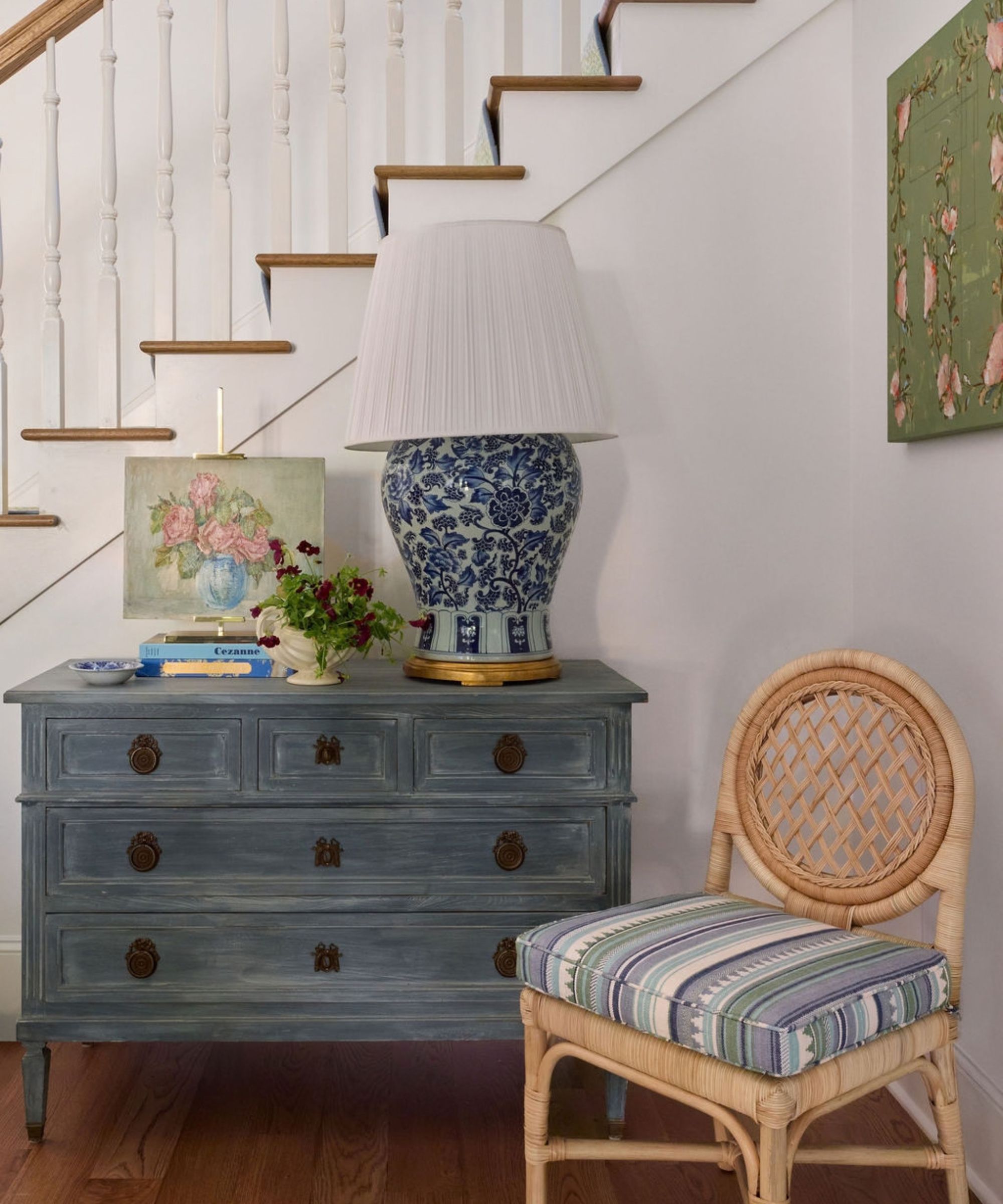 A distressed blue wooden dresser beneath a staircase, styled with a blue floral porcelain lamp and a woven rattan accent chair
