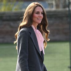 Kate Middleton walks outside wearing a grey coat with a pink blouse, grey trousers and grey high heels.