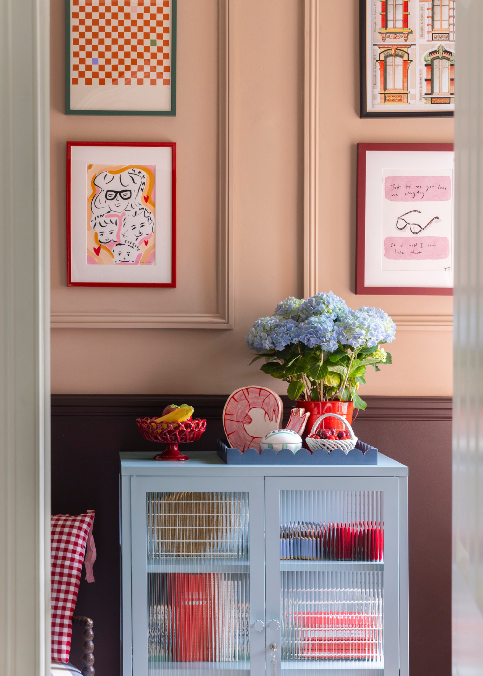 A room with dual color-drenched walls in cream and brown, featuring framed prints above and around. A blue fluted glass storage cabinet with a vase of hydrangeas and a bowl of items.