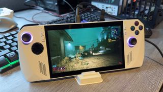A photo of an Asus ROG Ally handheld gaming PC, running Cyberpunk 2077, while rest in a white dock on a grey desk