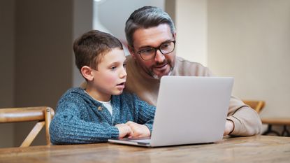 A young boy and his father look at a laptop together at a table.