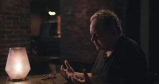 A man looks at his hands while sitting next to a table with a sculptural lamp, in a brick-walled room, in 'Look Into My Eyes.'