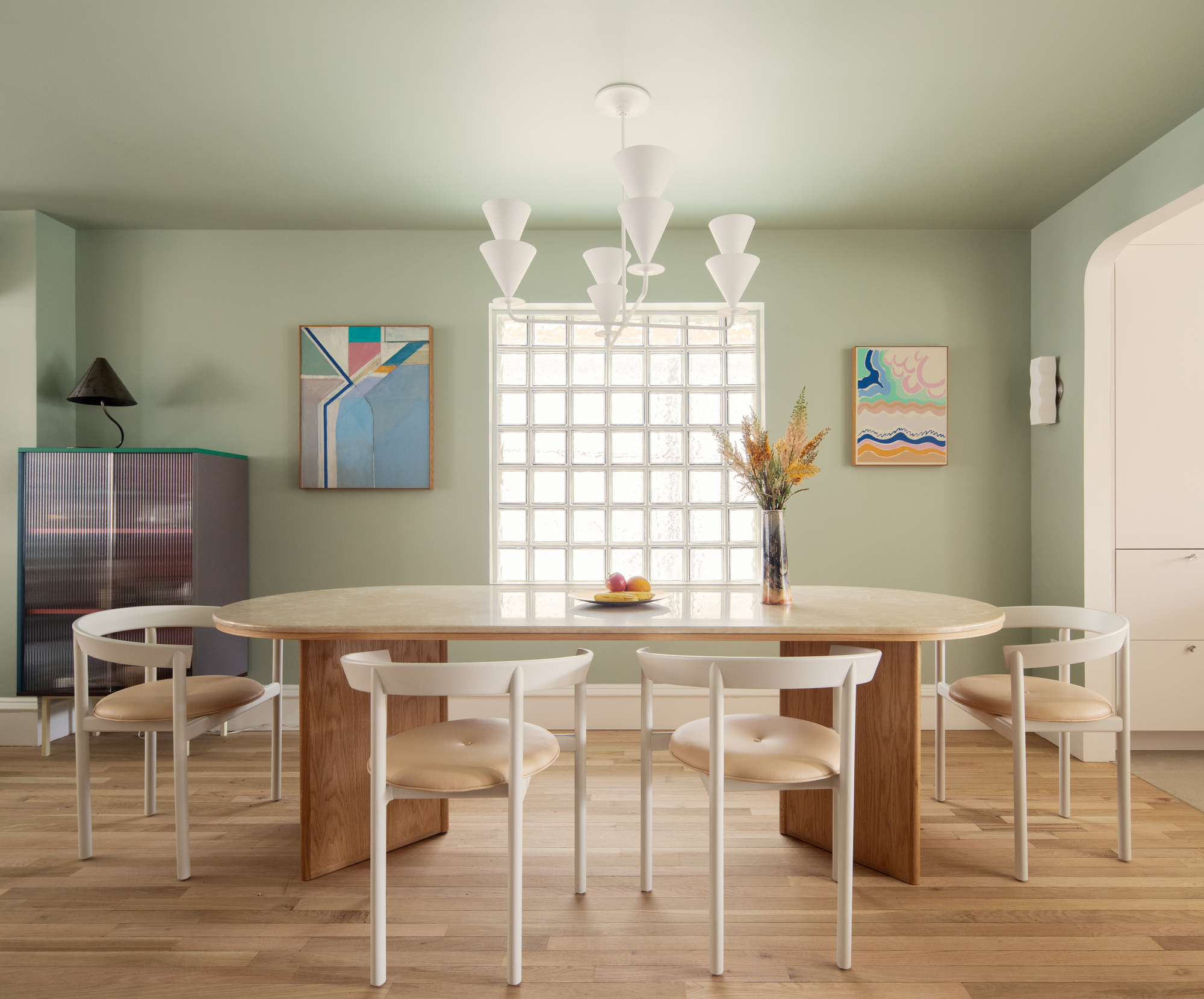 sage green dining room with glass block window, oval dining table with white chairs, artwork on the walls, and cabinet with lamp on top