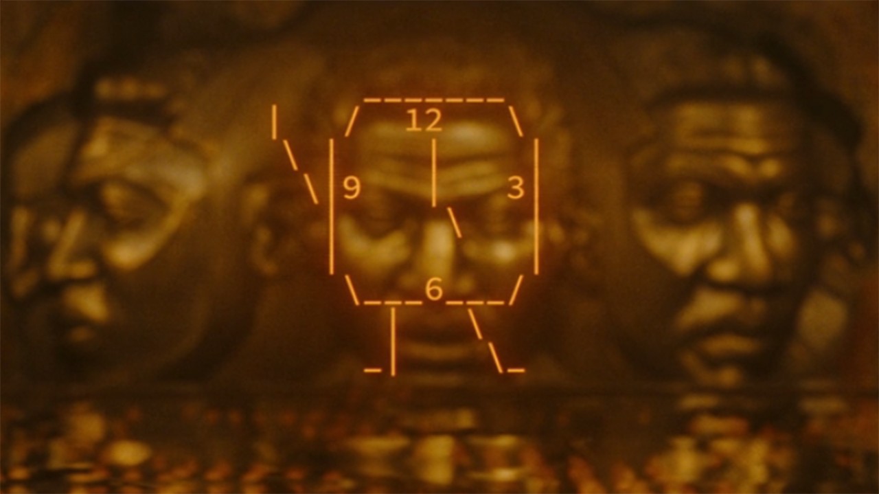 Image from the Marvel T.V. show Loki, season 2 episode 4. Here we see a cartoon clock made out of very basic orange lines showing the 12, 3, 6 and 9 positions on the clock. In the background there are three large golden faces.