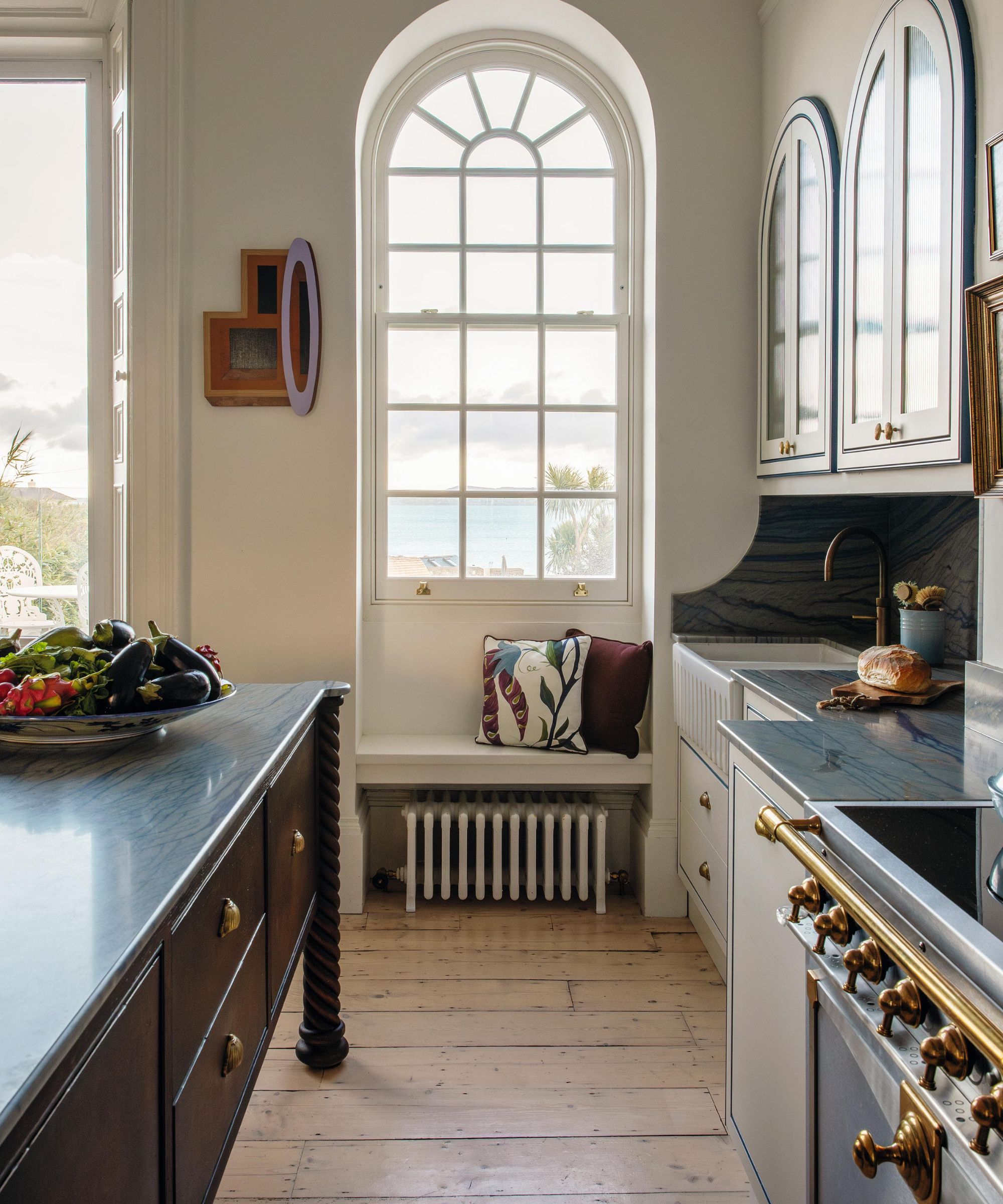 a white kitchen with arched windows overlooking the sea with a small window bench and arched kitchen cabinetry on the right. on the left, a dark wooden kitchen island