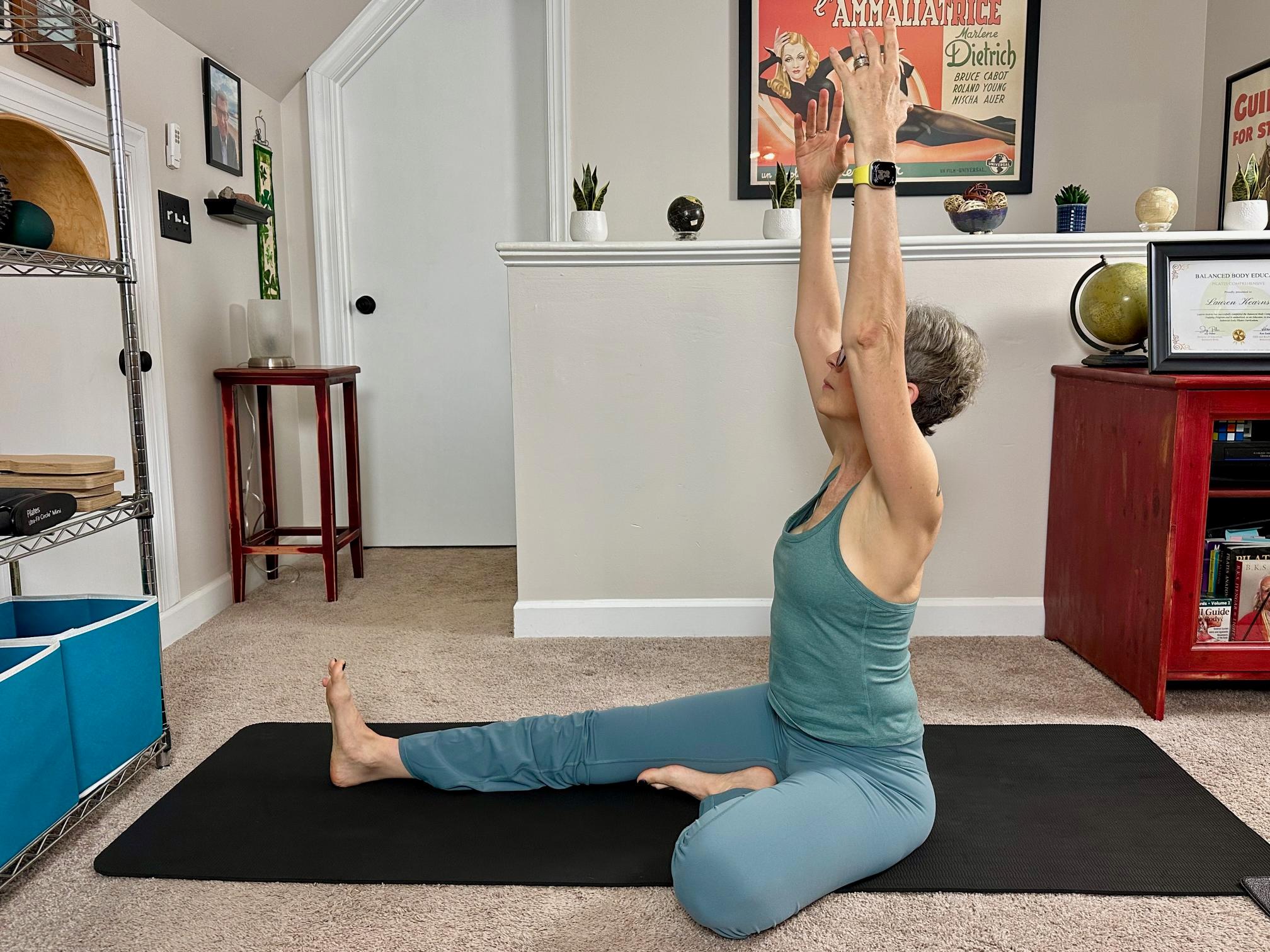 woman performing a yoga stretch sitting on the floor with one leg extended and the other bent in a living room on a black yoga mat