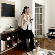 NYC fashion influencer Caroline Lin poses in her chic Manhattan living room wearing red lipstick, drop earrings, an open white button-down shirt, a neutral sweater over her shoulders, and black flared pants
