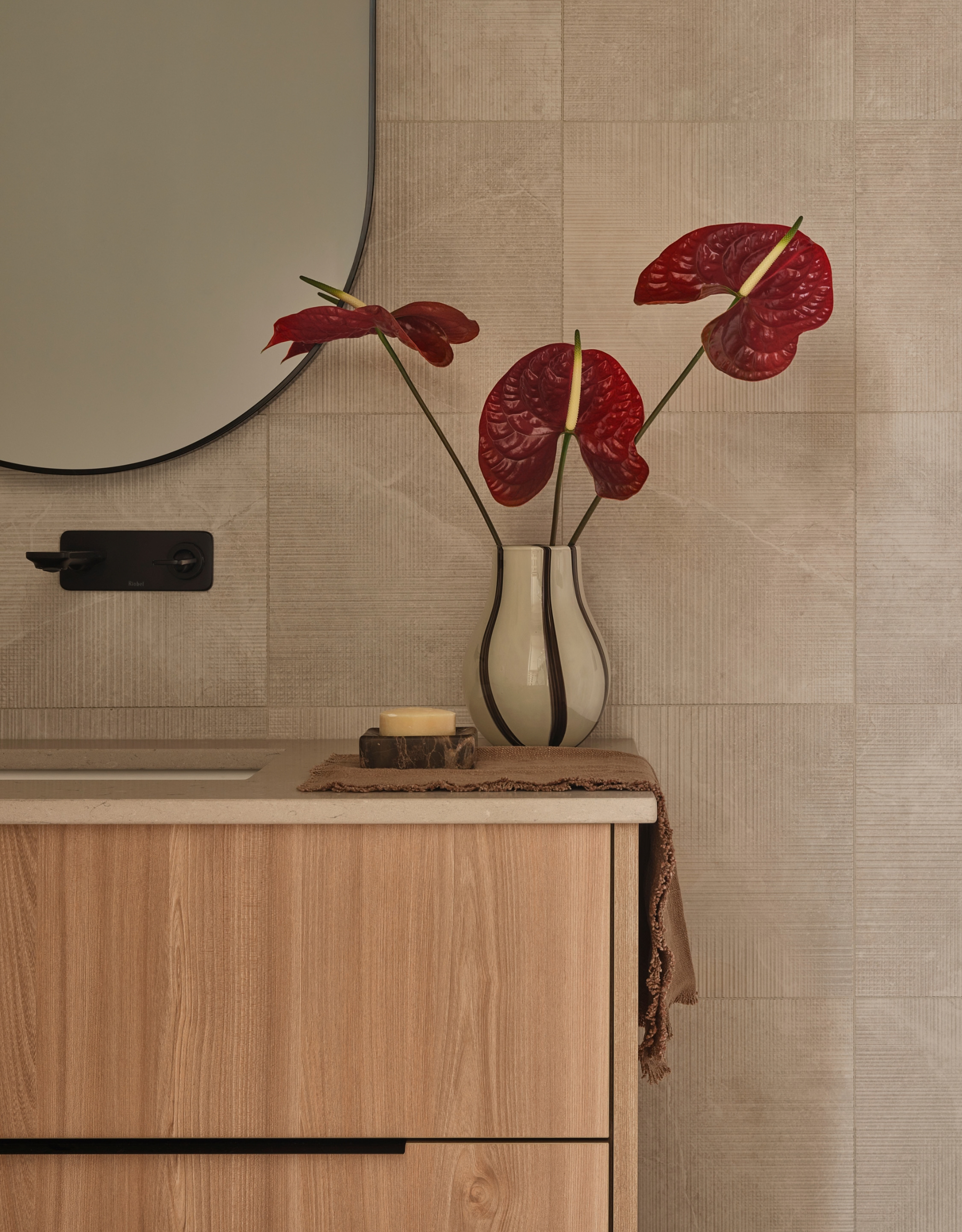 A minimalist bathroom with a floating wooden drawer and sink, a curved mirror, and a vase of red anthuriums