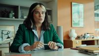 A dark-haired woman in a green blazer sits at an office desk, holding a small object between her fingers as she looks up mid-conversation.