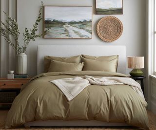 Sage green sheets and pillows on a bed against a white wall beneath landscape painting in wooden frames.