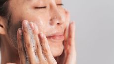 a woman close up washing her face with soap suds on her hands and a grey background