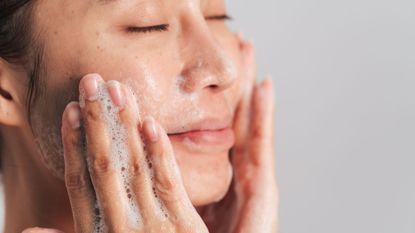 a woman close up washing her face with soap suds on her hands and a grey background