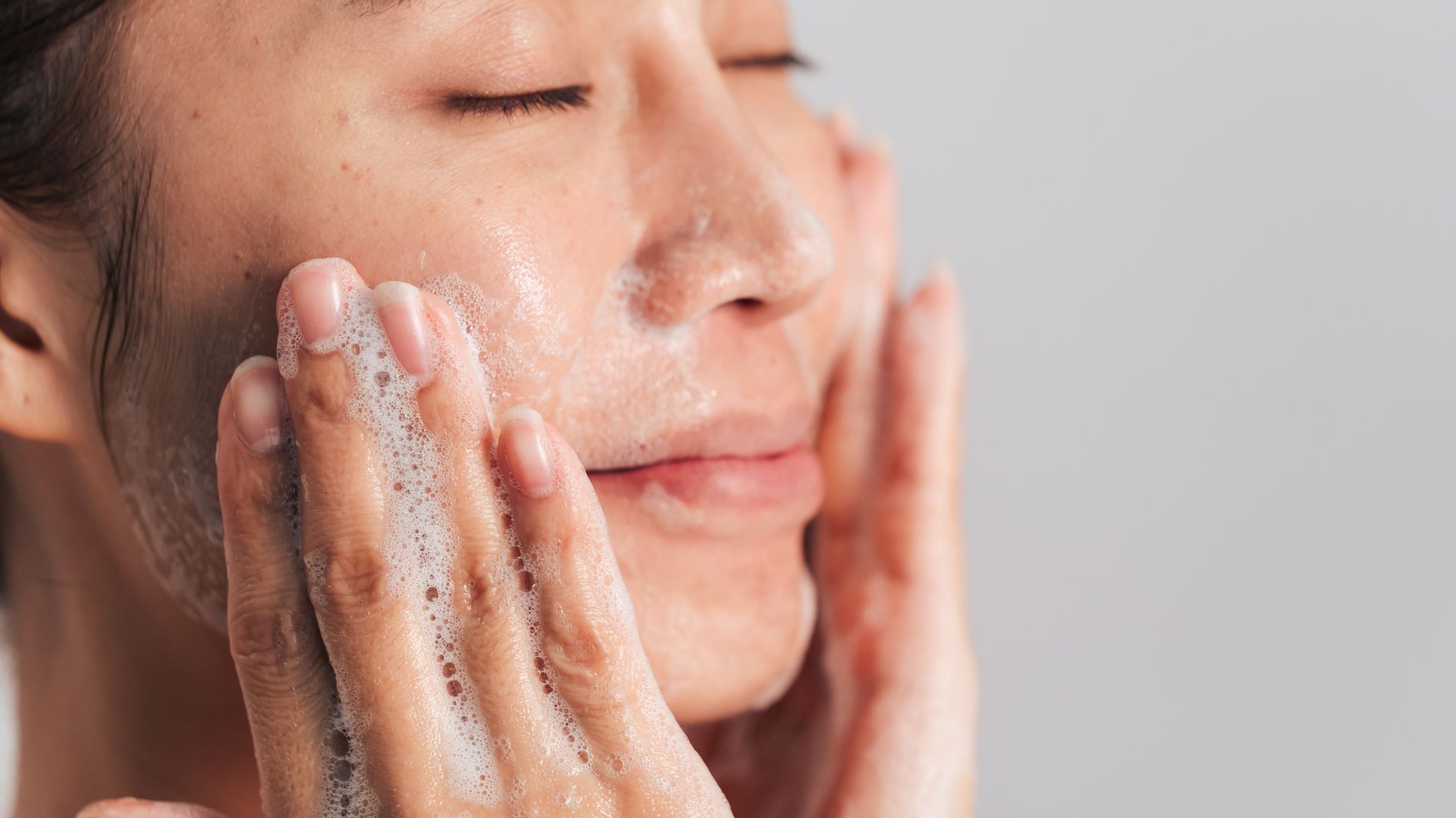 a woman close up washing her face with soap suds on her hands and a grey background