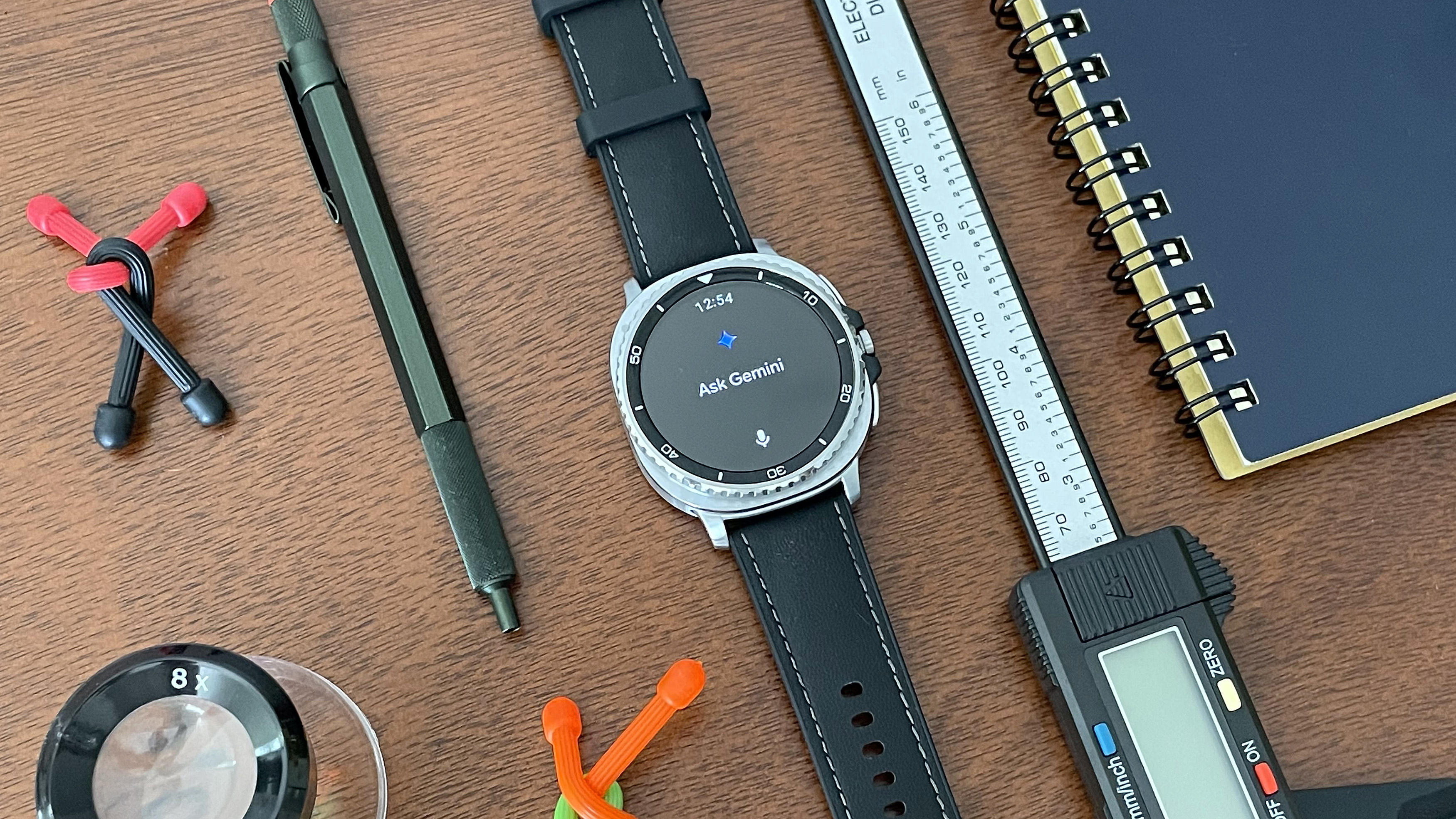 Close-up of the Samsung Galaxy Watch 8 Classic showing the Gemini AI assistant: the watch is on a wooden desk next to colorful items