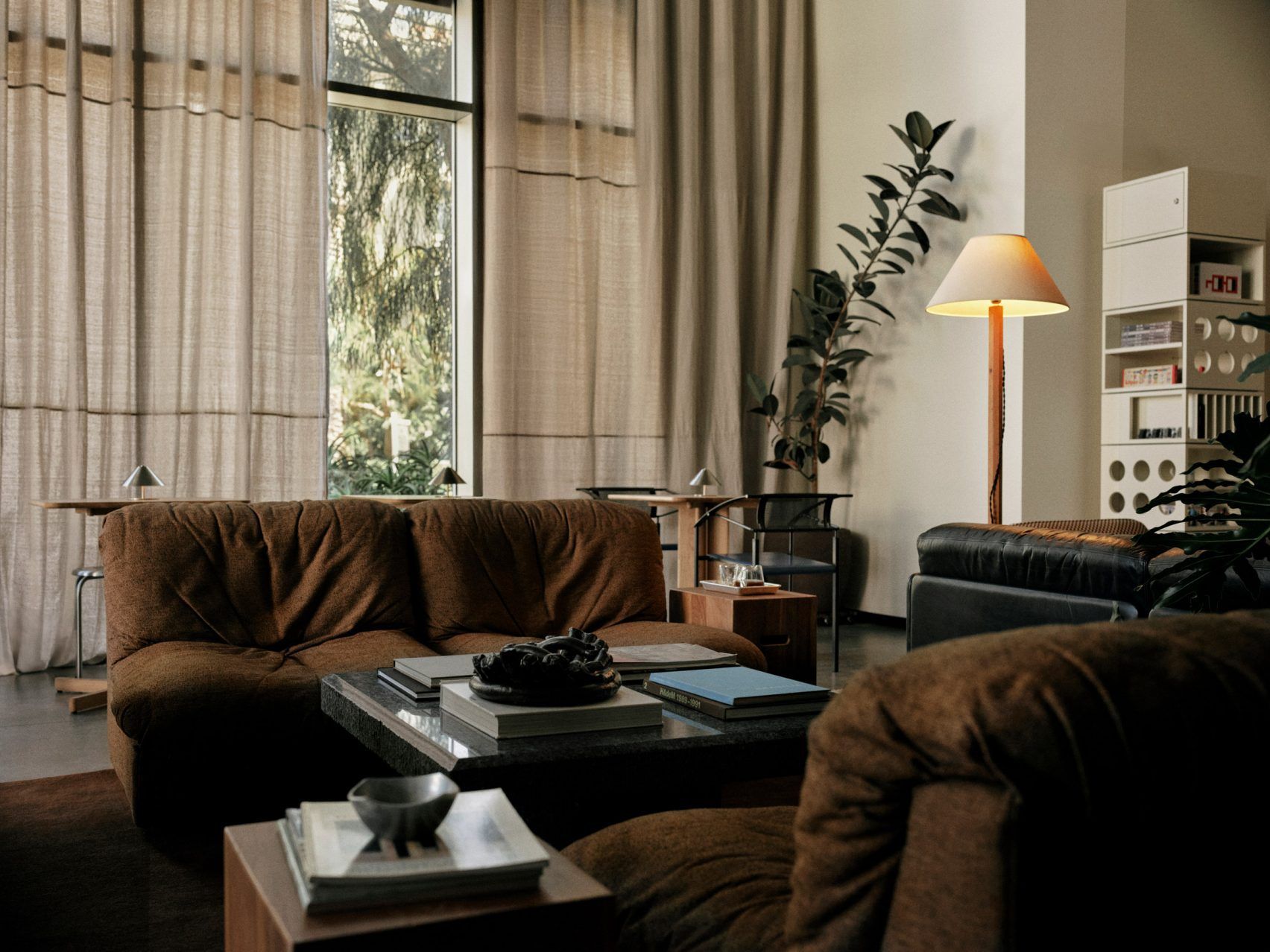 A home-looking coffee shop decorated with modernist furniture including spherical paper lanterns as lights, wood and chrome minimalist furniture, chrome table lamps, and thriving plants.