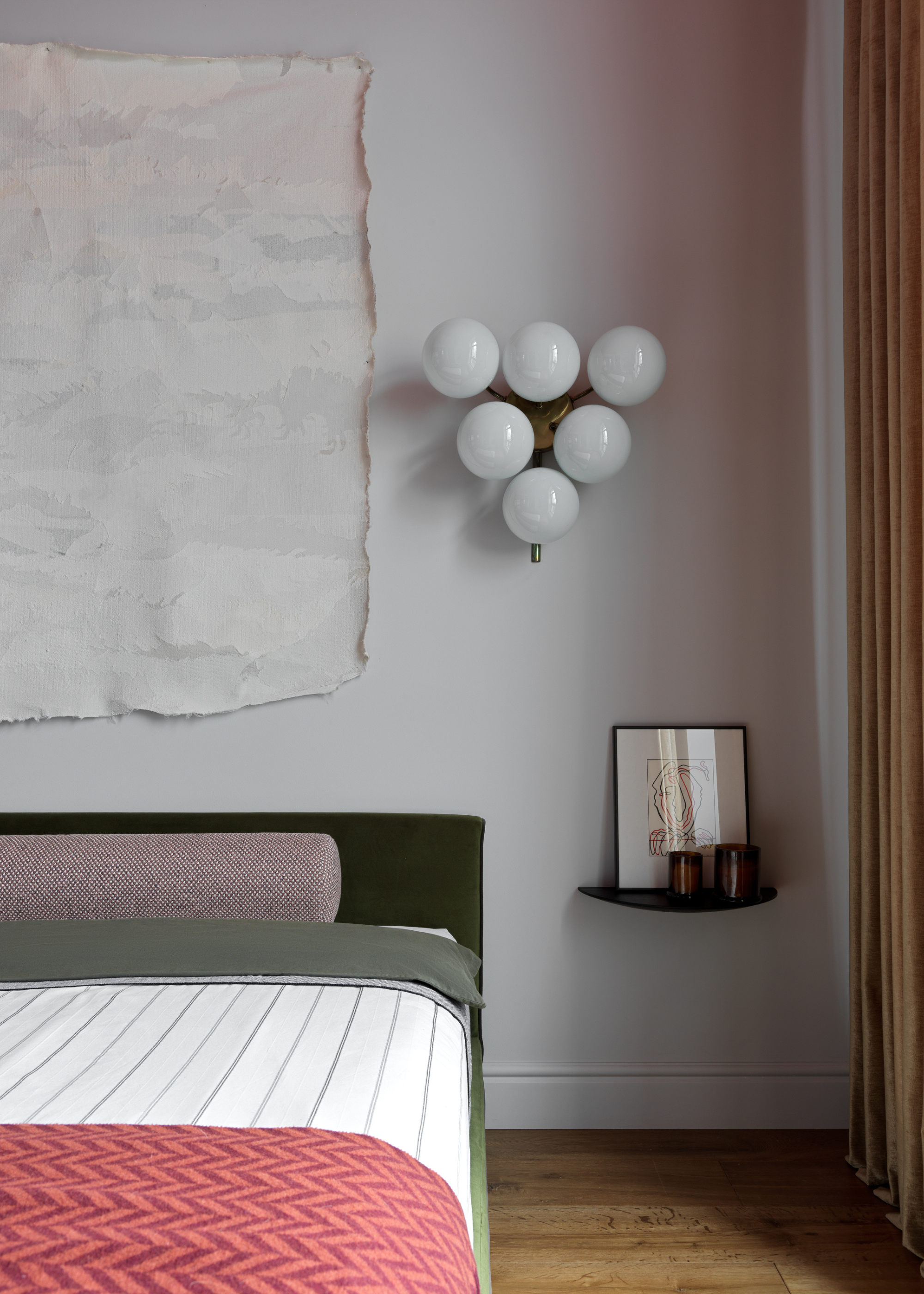 A white bedroom with a textured wall hanging, a maximalist wall sconce, a floating side table with amber glass candles and a leaning print, beside a green bed with a purple bolster pillow, a striped sheet and a colorful throw