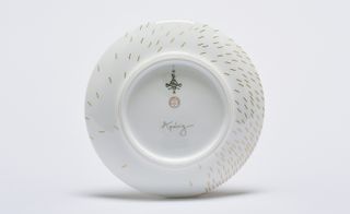 The underside of a white plate with its speckles continuing around the bottom edges. In the centre is the signature for Apeloig and a logo above.