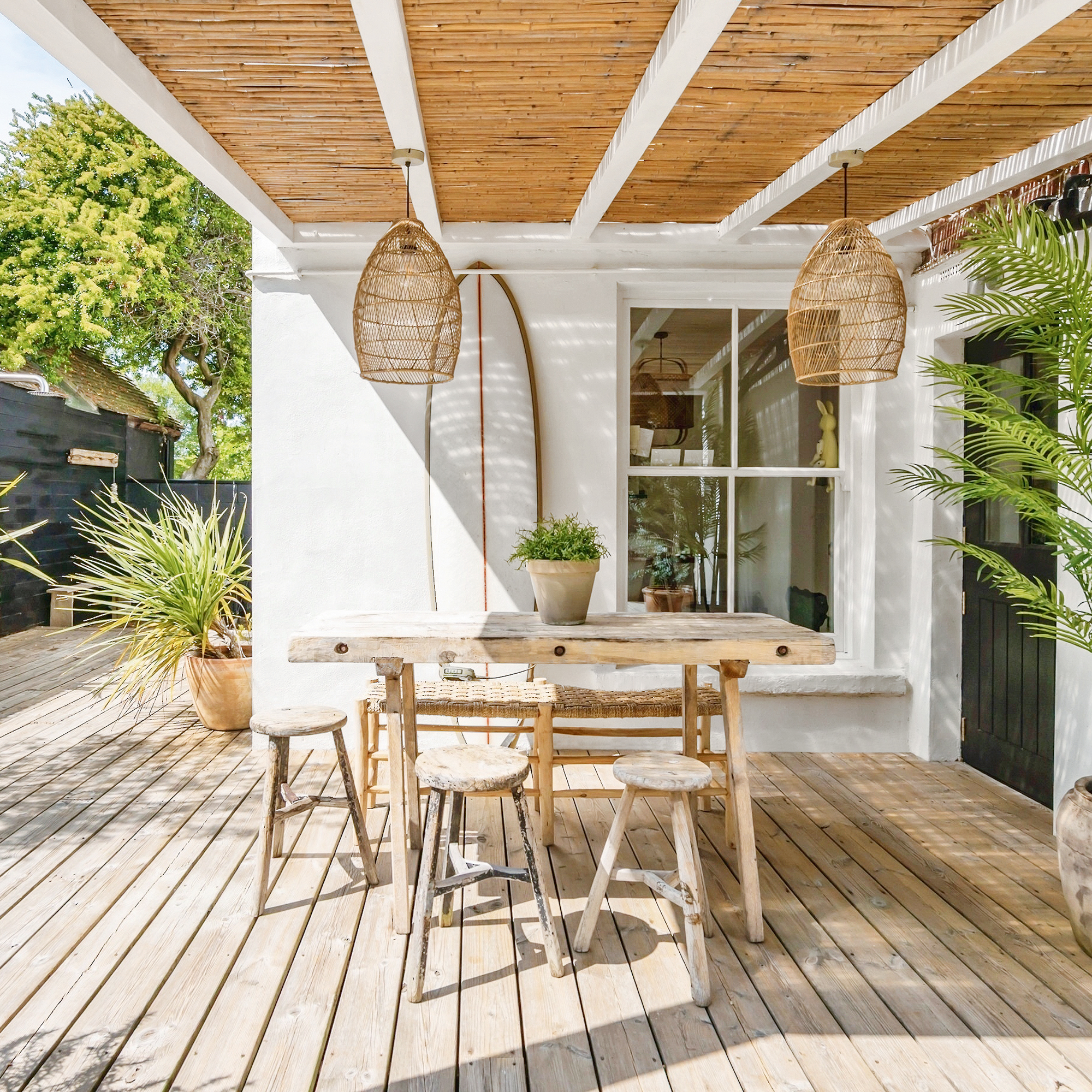 a sun drenched decked terrace with white walls and a surfboard, wicker pendant shades and a bamboo roof