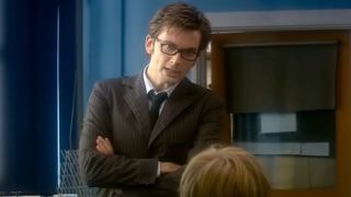 The 10th Doctor standing in front of a class with his arms crossed across his chest.