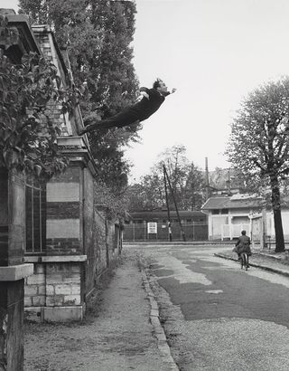 Black and white daytime image, uneven concrete road, man on a bicycle, stone brick building to the left, man in a dark suit jumping off the building wall, grey sky
