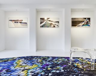 There are three abstract paintings on the far wall. On the floor, there is a blue-toned rug in geometrical shapes, with a see-through stool sitting on it.